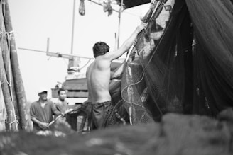 A group of men are engaged in handling large fishing nets. One man, shirtless, is in focus as he lifts a section of the net. Others in the background appear to be helping or observing.