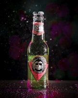 A close-up of a glass bottle with the label 'Icy Monkey Natural Boost'. The bottle is green and appears to be chilled with condensation droplets on its surface. The label features an illustration of a monkey wearing sunglasses. The background is dark with a bokeh effect, featuring hints of purple and pink lights.
