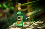 A green glass bottle of Loy Monkey drink sits on a surface with blurred green leaves in the background, giving a sense of an outdoor garden or natural setting. The label on the bottle features a cartoon monkey wearing sunglasses and text in both English and Arabic.