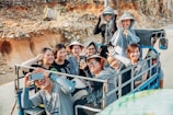 A group of people sitting closely together on a small vehicle, smiling and taking photos. They are wearing casual clothing and some have hats or scarves. The background shows a rocky landscape, indicating they might be on a trip or adventure.