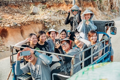 A group of people sitting closely together on a small vehicle, smiling and taking photos. They are wearing casual clothing and some have hats or scarves. The background shows a rocky landscape, indicating they might be on a trip or adventure.