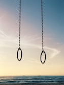 Two gymnastic rings hang from chains against a serene beach backdrop. The sky transitions from soft pastel hues to deeper blues, and the ocean extends towards the horizon with gentle waves.