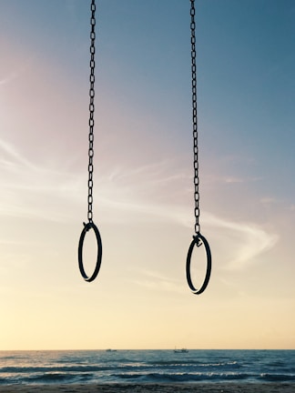 Two gymnastic rings hang from chains against a serene beach backdrop. The sky transitions from soft pastel hues to deeper blues, and the ocean extends towards the horizon with gentle waves.