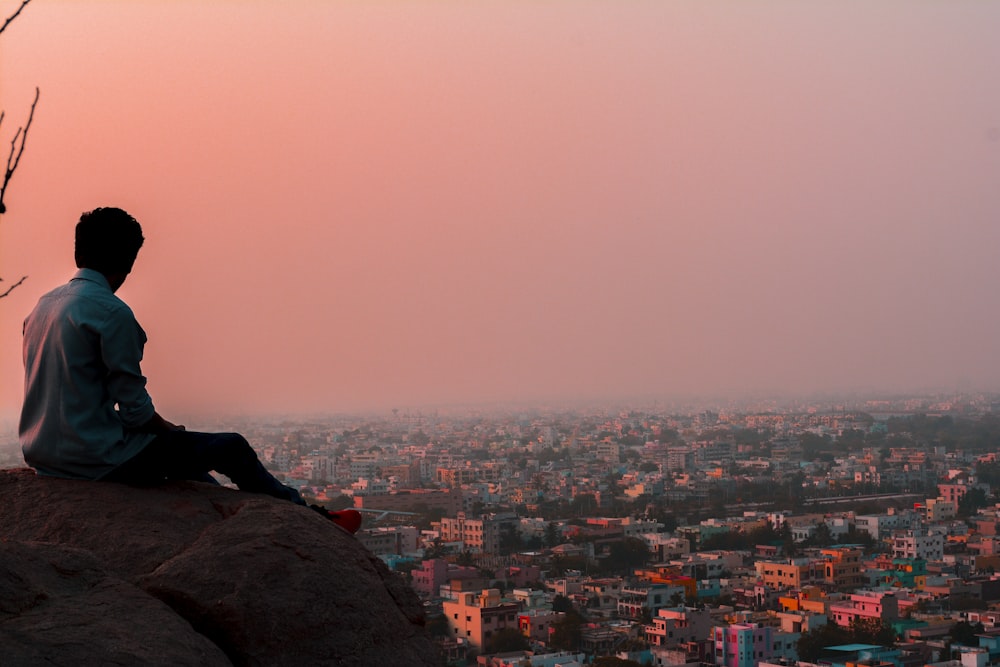 man sitting on rock formation facing on buildings during daytime