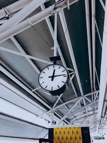 A large analog clock is suspended from the ceiling of a modern architectural structure with metal beams and panels. The clock features bold black hands and markings on a white face. Below the clock, there is a decorative yellow and black patterned element, possibly part of a sign or information board.