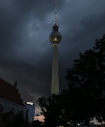 A tall television tower stands prominently against a dramatic, cloudy sky. The tower is illuminated, with multiple small red lights visible along its structure. Adjacent buildings and trees are in silhouette, adding to the moody atmosphere.