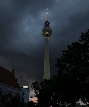 A tall television tower stands prominently against a dramatic, cloudy sky. The tower is illuminated, with multiple small red lights visible along its structure. Adjacent buildings and trees are in silhouette, adding to the moody atmosphere.