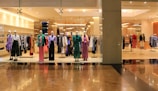 Mannequins dressed in various colorful outfits are displayed in a fashion store window. The interior is brightly lit, showcasing racks filled with neatly organized clothing items. The store has a modern, luxurious appearance with polished flooring.