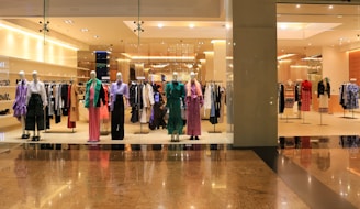 Mannequins dressed in various colorful outfits are displayed in a fashion store window. The interior is brightly lit, showcasing racks filled with neatly organized clothing items. The store has a modern, luxurious appearance with polished flooring.
