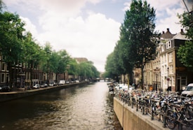 A picturesque canal scene lined with traditional European buildings and lush green trees on either side. Numerous bicycles are parked along the canal's edge, illustrating a common mode of transportation in this region. The sky is partly cloudy, casting a gentle light over the scene, contributing to a serene atmosphere.