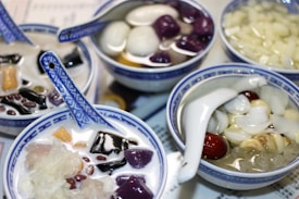 Several bowls of Asian desserts, each filled with a variety of ingredients like taro balls, red beans, black jelly, and other sweet ingredients in a syrupy broth. The bowls are made of porcelain, decorated with intricate blue patterns, and each contains a matching blue spoon.