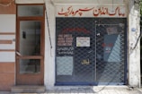 A storefront with a closed metal security grille covering the entrance. The sign above the door has text in a foreign script, and additional information is displayed on the glass door. The facade is simple, with a combination of tiles and plain walls.