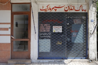 A storefront with a closed metal security grille covering the entrance. The sign above the door has text in a foreign script, and additional information is displayed on the glass door. The facade is simple, with a combination of tiles and plain walls.