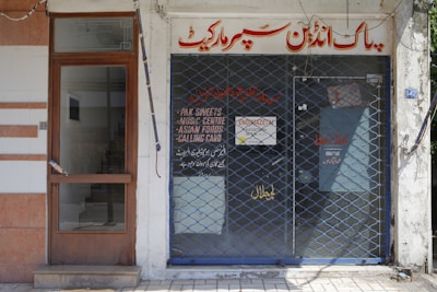 A storefront with a closed metal security grille covering the entrance. The sign above the door has text in a foreign script, and additional information is displayed on the glass door. The facade is simple, with a combination of tiles and plain walls.