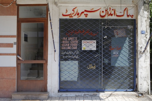 A storefront with a closed metal security grille covering the entrance. The sign above the door has text in a foreign script, and additional information is displayed on the glass door. The facade is simple, with a combination of tiles and plain walls.