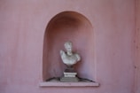 A classical marble bust is placed in an arched niche within a pink stucco wall. The bust is of a man with curly hair, displayed prominently, while the wall shows signs of wear with small dark spots.