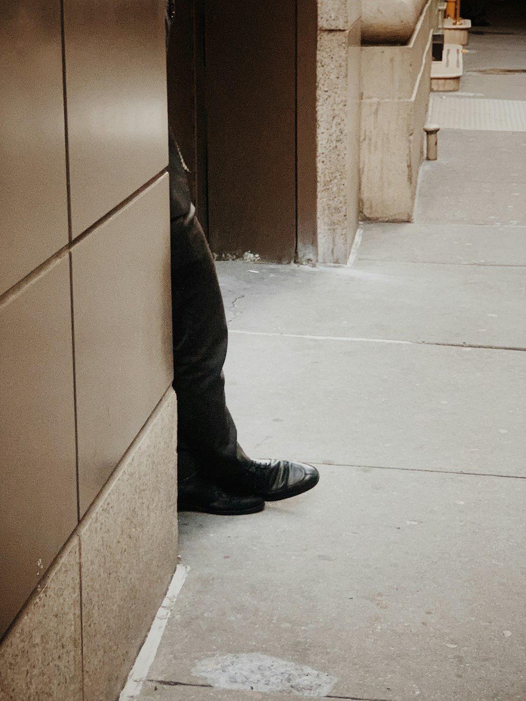 man wearing black pants and leather shoes leaning near brown wall