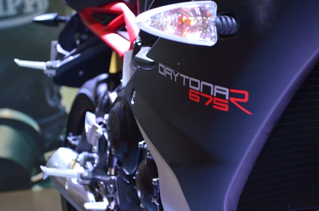 Close-up view of a motorcycle with the brand name 'Daytona 675R' prominently displayed in bold lettering. The motorcycle has a sleek design with a black and red color scheme, emphasizing its sporty appearance. A visible part of the engine and a rear-view mirror can be seen, along with foot pegs and a metallic exhaust pipe.