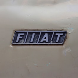 A close-up of a metallic emblem with the word 'FIAT' embossed on it. The emblem is slightly worn with visible scratches and signs of age. The background is a textured, pale beige surface that appears to be part of a vehicle’s body.