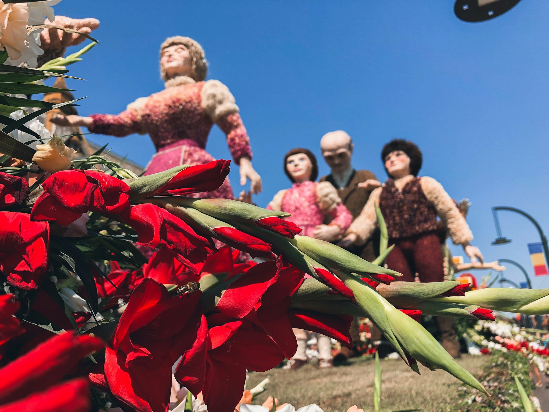 A vibrant scene featuring red gladiolus flowers in the foreground, with four life-sized human figures constructed from flowers in the background. The sky is clear and blue, adding to the colorful ambiance.