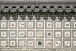 A traditional wall decorated with tiles, each featuring different carved images including various animals, plants, and abstract designs. The roof above the wall has intricately designed tiles with a circular pattern.