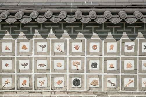 A traditional wall decorated with tiles, each featuring different carved images including various animals, plants, and abstract designs. The roof above the wall has intricately designed tiles with a circular pattern.