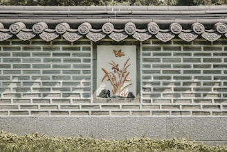 A traditional stone and brick wall features ornate decorative patterns along the roofline. A central panel displays an artistic depiction of a bird above bamboo plants in relief. Shadows play across the structure, adding depth to the intricate details.
