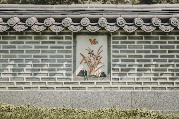 A traditional stone and brick wall features ornate decorative patterns along the roofline. A central panel displays an artistic depiction of a bird above bamboo plants in relief. Shadows play across the structure, adding depth to the intricate details.