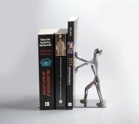 Three books are aligned vertically next to a metal bookend shaped like a human figure, which appears to be pushing or holding the books upright. The bookend is metallic and reflective.