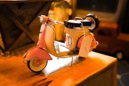A small, intricately detailed model of a vintage pink and white scooter is placed on a wooden surface. The scooter features realistic components like a headlight, handlebars, and a seat. Attached to the scooter is a price tag indicating it is for sale. In the background, there are blurred objects that appear to be a wooden crate and possibly an old television set, creating a nostalgic atmosphere.