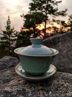 A green ceramic tea cup with a lid is placed on a rock, set against the backdrop of trees and a sunset sky. The scene is captured outdoors, blending natural elements with the handcrafted pottery piece.