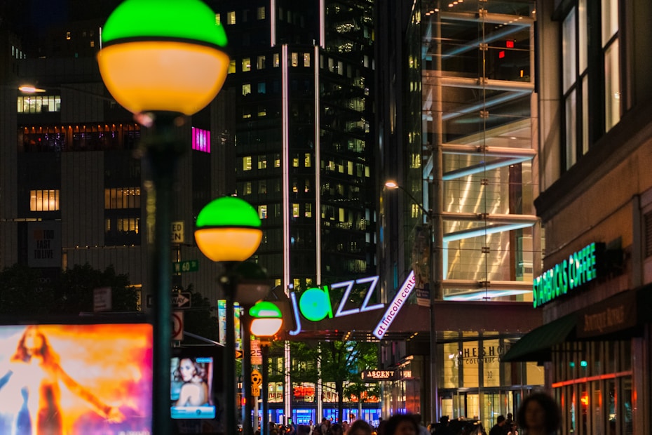 City street at night illuminated by bright signs and streetlights. A prominent jazz club sign is visible, with a busy city backdrop. Green and orange glowing subway entrance lights add to the urban atmosphere.
