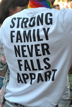 A person is wearing a white T-shirt with bold black text that reads 'STRONG FAMILY NEVER FALLS APPART'. The individual has a colorful, striped scarf around their neck, suggesting a possible pride-related event. Part of another person is visible to the left, and there are blurred people in the background.