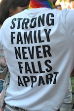 A person is wearing a white T-shirt with bold black text that reads 'STRONG FAMILY NEVER FALLS APPART'. The individual has a colorful, striped scarf around their neck, suggesting a possible pride-related event. Part of another person is visible to the left, and there are blurred people in the background.