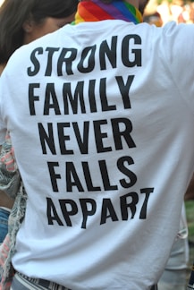 A person is wearing a white T-shirt with bold black text that reads 'STRONG FAMILY NEVER FALLS APPART'. The individual has a colorful, striped scarf around their neck, suggesting a possible pride-related event. Part of another person is visible to the left, and there are blurred people in the background.