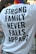 A person is wearing a white T-shirt with bold black text that reads 'STRONG FAMILY NEVER FALLS APPART'. The individual has a colorful, striped scarf around their neck, suggesting a possible pride-related event. Part of another person is visible to the left, and there are blurred people in the background.