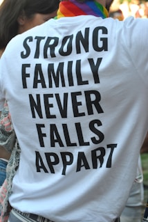 A person is wearing a white T-shirt with bold black text that reads 'STRONG FAMILY NEVER FALLS APPART'. The individual has a colorful, striped scarf around their neck, suggesting a possible pride-related event. Part of another person is visible to the left, and there are blurred people in the background.