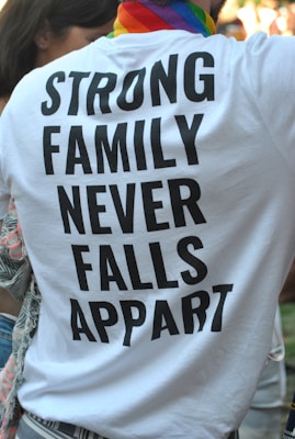 A person is wearing a white T-shirt with bold black text that reads 'STRONG FAMILY NEVER FALLS APPART'. The individual has a colorful, striped scarf around their neck, suggesting a possible pride-related event. Part of another person is visible to the left, and there are blurred people in the background.