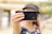 A person is holding a smartphone in front of their face, partially obscuring their features. The focus is on the phone, with a blurred background. The hand holding the phone is adorned with gold bracelets.