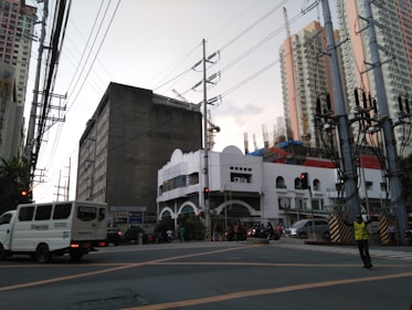 A busy urban intersection with several tall buildings in the background. Power lines and a traffic light are visible. A jeepney and other vehicles are on the road, and a traffic enforcer stands at the corner. The sky is overcast, and construction activities can be seen in the distance.