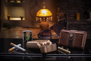 A collection of personal items is arranged on a reflective surface in a dimly lit room featuring a warm, ambient glow from a decorative lamp. The items include a pair of tortoiseshell sunglasses, a leather wallet, a wooden cigarette case with cigarettes, a metal lighter with a green design, and a small cylindrical object. The background includes shelves with small potted plants and decorative elements.