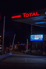A dimly lit gas station at night with the 'Total' signage prominently glowing in red at the top. Below, a garage or service section is indicated by a sign which reads 'Mecanique Carrosserie.' The surrounding area is dark, with some visible parked cars and a few streetlights casting a faint glow.