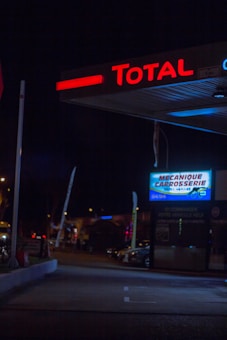A dimly lit gas station at night with the 'Total' signage prominently glowing in red at the top. Below, a garage or service section is indicated by a sign which reads 'Mecanique Carrosserie.' The surrounding area is dark, with some visible parked cars and a few streetlights casting a faint glow.