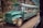 A vintage green bus is traveling on a narrow, winding road in a mountainous area. The bus is filled with passengers looking out from the windows, creating a sense of adventure. The surrounding environment includes rocky cliffs and dense greenery, adding to the scenic and rugged atmosphere.