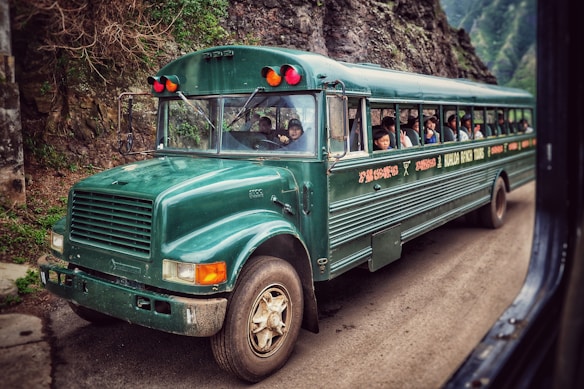 A vintage green bus is traveling on a narrow, winding road in a mountainous area. The bus is filled with passengers looking out from the windows, creating a sense of adventure. The surrounding environment includes rocky cliffs and dense greenery, adding to the scenic and rugged atmosphere.