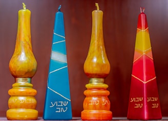 Four decorative candles with Hebrew inscriptions are displayed against a plain background. The candles vary in color and shape, including shades of orange, blue, yellow, and red. Two of the candles have a tear-shaped form, while the others are pyramid-shaped.