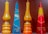 Four decorative candles with Hebrew inscriptions are displayed against a plain background. The candles vary in color and shape, including shades of orange, blue, yellow, and red. Two of the candles have a tear-shaped form, while the others are pyramid-shaped.