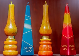 Four decorative candles with Hebrew inscriptions are displayed against a plain background. The candles vary in color and shape, including shades of orange, blue, yellow, and red. Two of the candles have a tear-shaped form, while the others are pyramid-shaped.