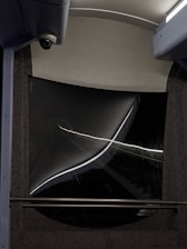 A dark tunnel view captured from the rear window of a vehicle, with strong reflections and light streaks creating an abstract effect. The window frame has a curved, futuristic design with a security camera visible on the top left corner.