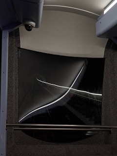 A dark tunnel view captured from the rear window of a vehicle, with strong reflections and light streaks creating an abstract effect. The window frame has a curved, futuristic design with a security camera visible on the top left corner.